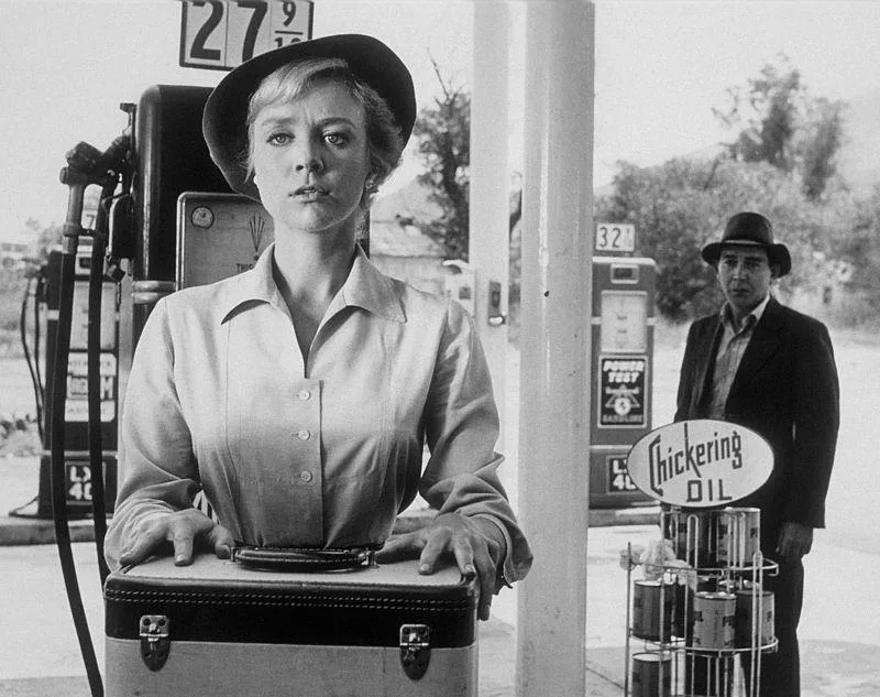 A woman resembling Inger Stevens standing at a gas station with a suitcase.