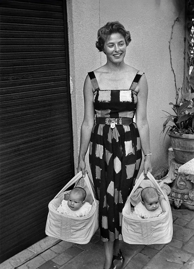 Ingrid Bergman in a summer dress holding her twins in two baby baskets in each hand.