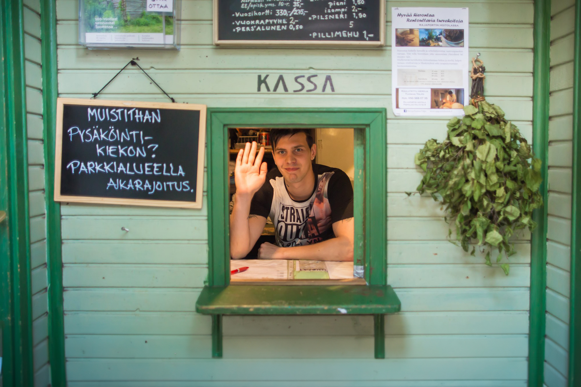 A man waves from a small green cashier window. Beside him, a chalkboard with Finnish text hangs on the wall, and a bunch of green herbs is attached to the wood paneling.
