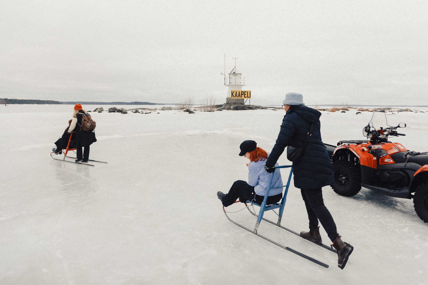 Three people are on an ice-covered surface; two are using kicksleds while one person is being pushed on a sled by another. A red all-terrain vehicle is parked nearby, with a small structure visible in the distance, enjoying kick sledding over Frozen Lake Näsijärvi near Siilinkari Light House in Tampere.