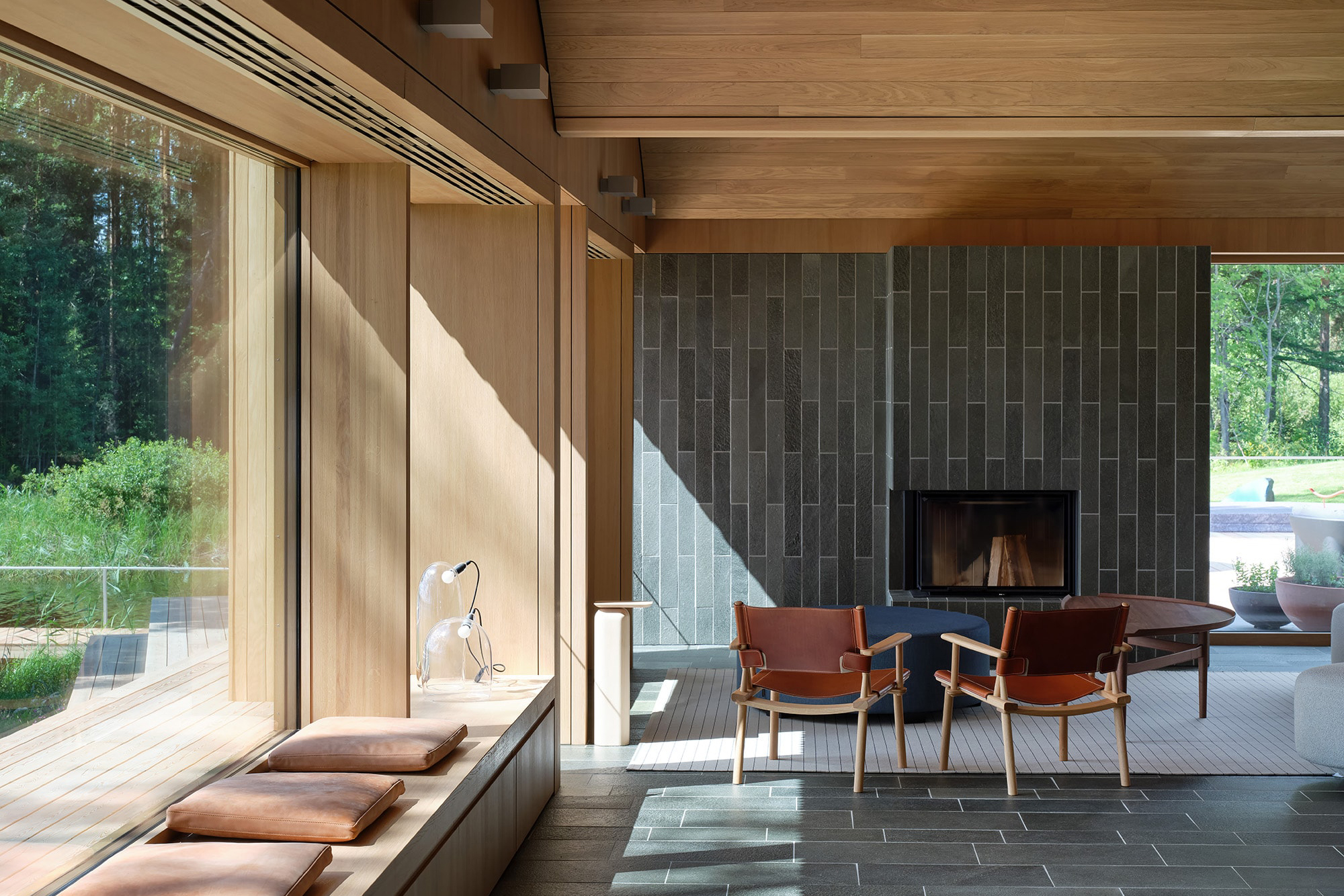 A modern living room with wood-paneled walls and large windows, reminiscent of a Serlachius Art Sauna. It features a fireplace, chairs, and a low table. Sunlight streams in, highlighting the minimalist design and greenery outside.