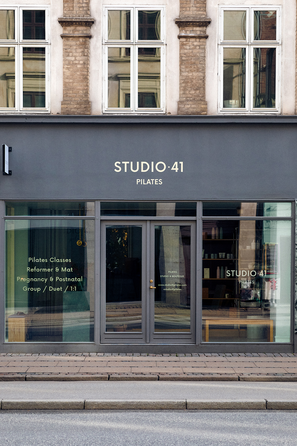 Storefront view of Studio 41 Pilates with large windows displaying class offerings including group, duet, and one-on-one sessions. Brick facade with gray signage above the entrance.