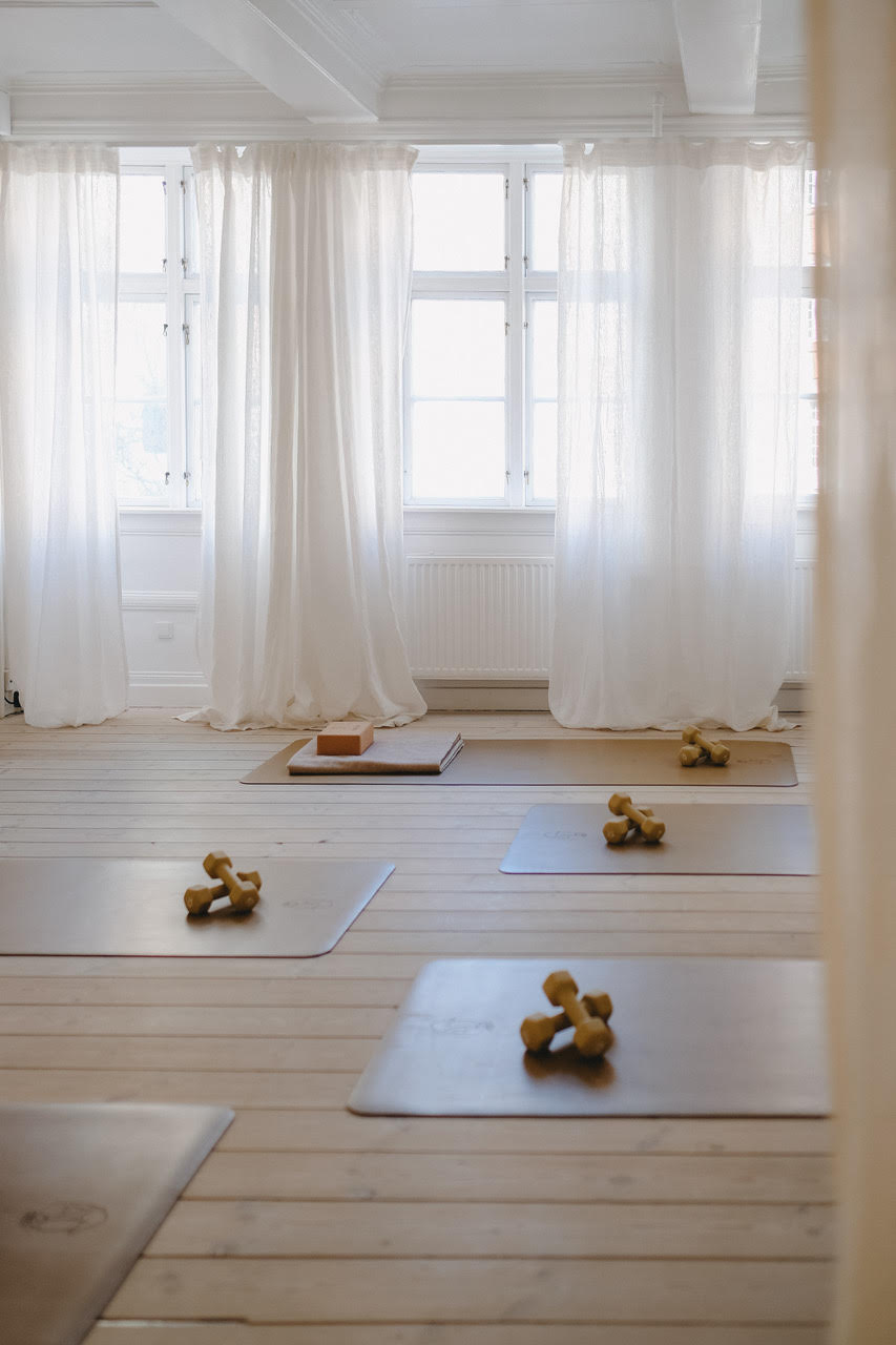 A bright studio with yoga mats, small dumbbells, and yoga blocks arranged on a light wooden floor near large windows with sheer curtains.