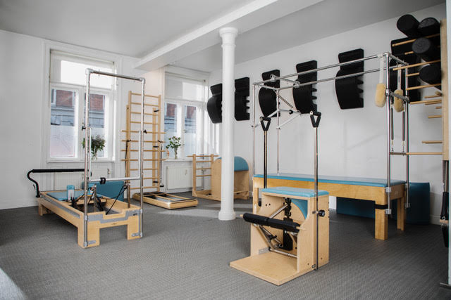 A bright, minimalist Pilates studio featuring various reformer and trapeze equipment, wall bars, and padded exercise areas on a grey carpeted floor.